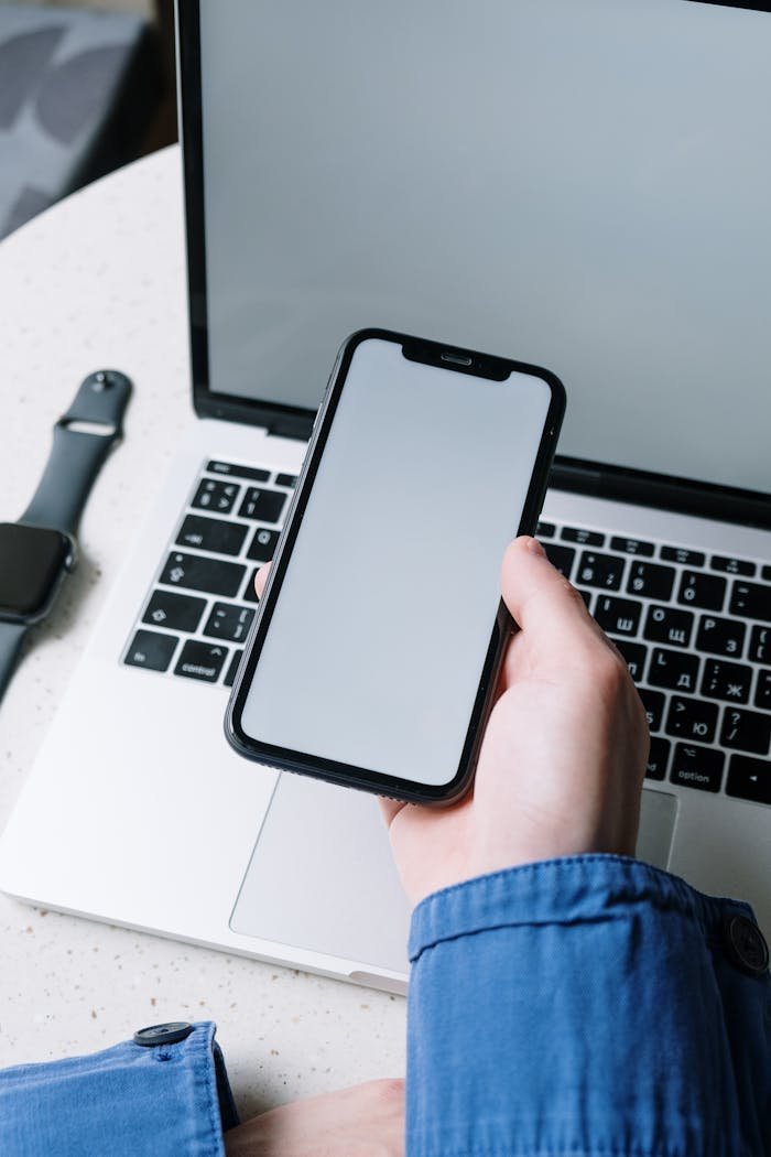 services-02 A hand holding a smartphone with a blank screen above a laptop, showcasing modern technology.